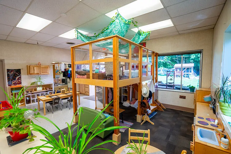 The image displays an indoor play area at Chemeketa's Child Development Center. At the center of the room is a large wooden structure resembling a fortress with multiple levels. To the left, there are tables and chairs arranged around a counter with various items, including books, toys, and a potted plant. On the right side, there is a window